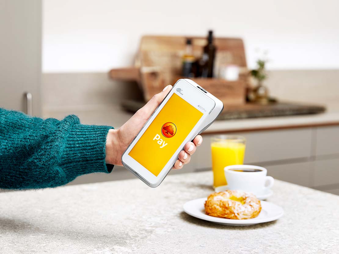 The image shows someone seated in a cafe with a glass of juice, a cup of coffee and a cake on the table in front of them, using a card terminal to pay.